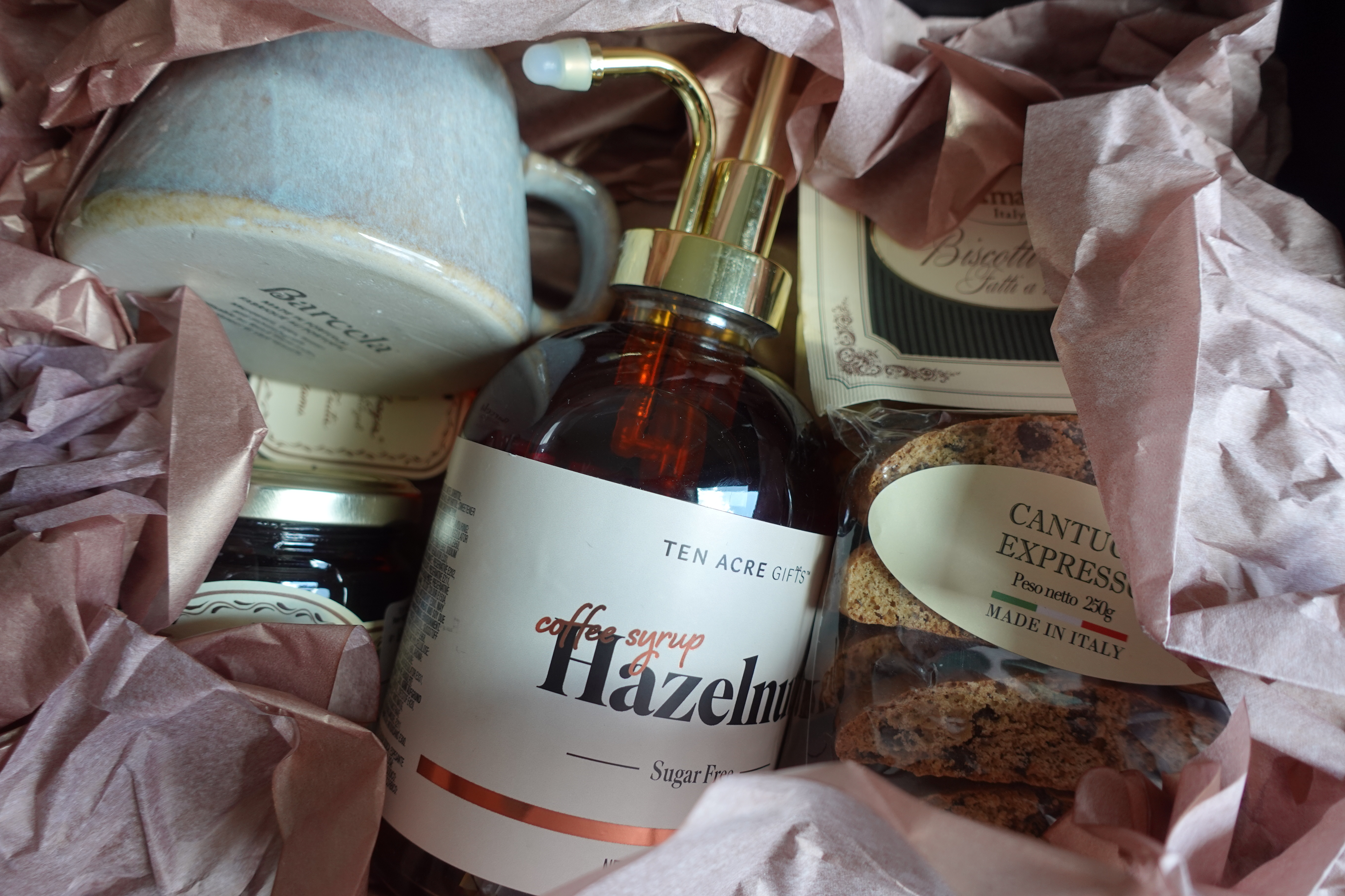 Close-up of a mug, coffee syrup, and biscotti nestled in rose tissue paper
