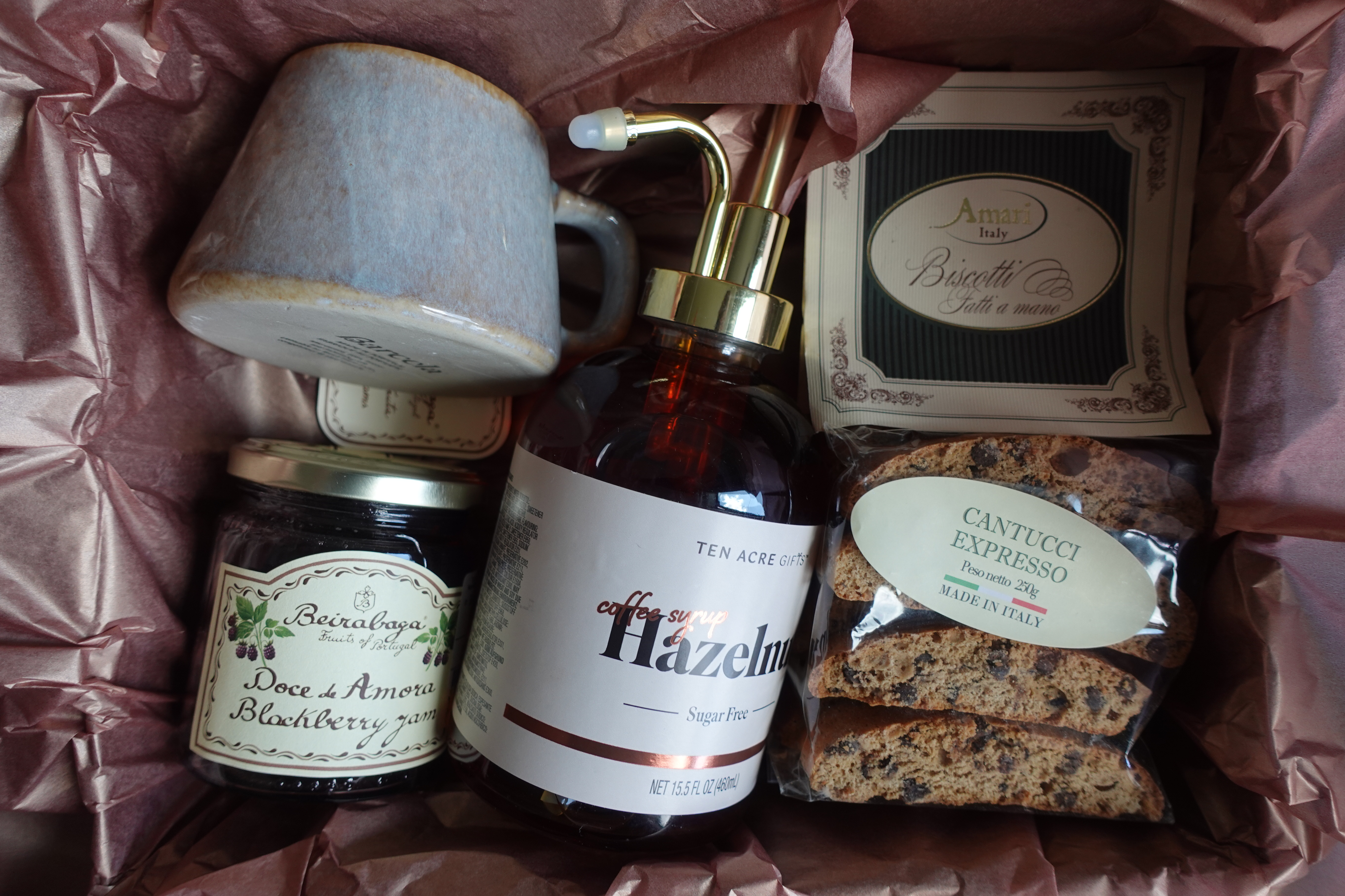 A curated gift box opened to reveal a mug, coffee syrup, jam, and biscotti wrapped in rose tissue paper
